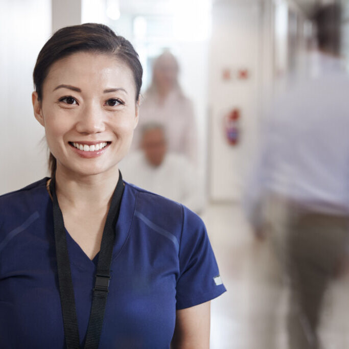 Portrait Of Smiling Female Nurse Wearing Scrubs In Busy Hospital Corridor