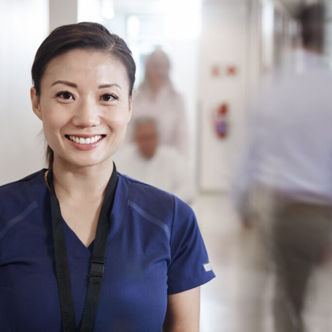 Portrait Of Smiling Female Nurse Wearing Scrubs In Busy Hospital Corridor Portrait Of Smiling Female Nurse Wearing Scrubs In Busy Hospital Corridor